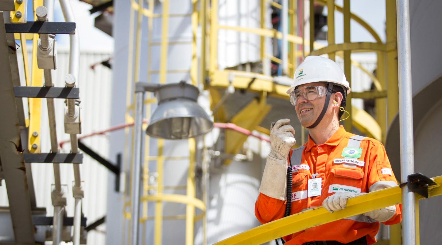 Petrobras professional wearing an orange uniform and PPE communicating by radio in an industrial area.