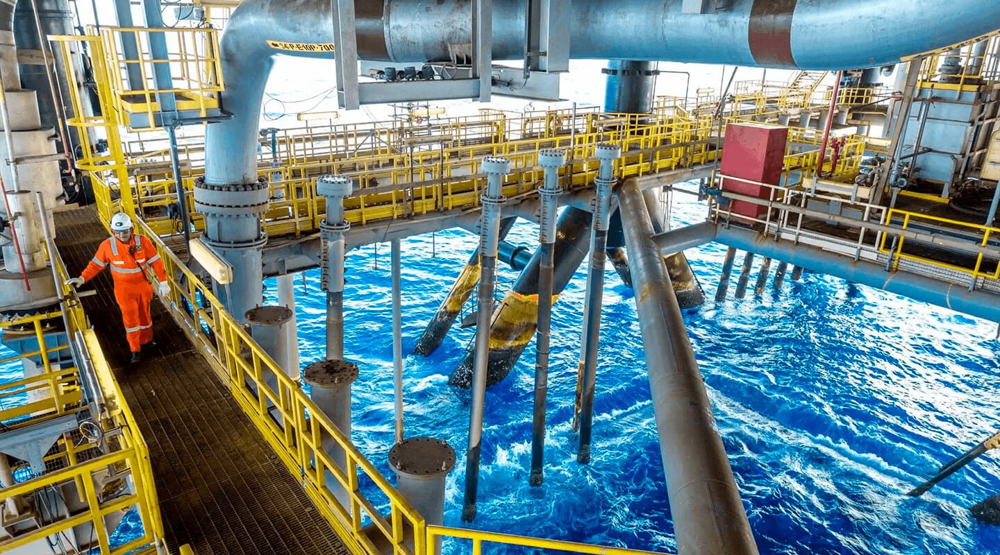 Offshore oil platform worker in orange protective uniform walking along metal walkway above the ocean.
